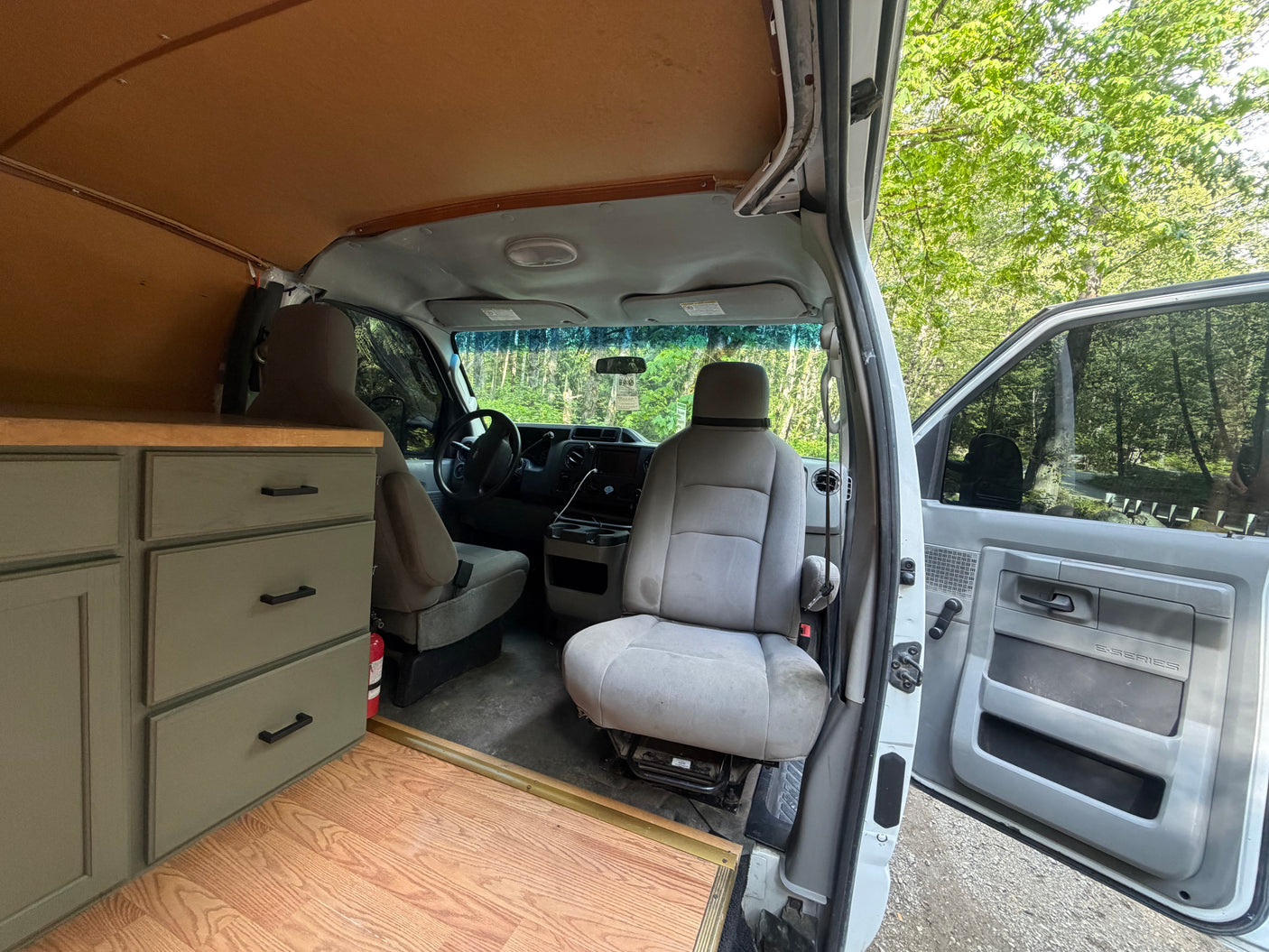 Inside this custom RV rental camper van: light brown ceiling, grey cabinets, front seats, and a view of lush trees.