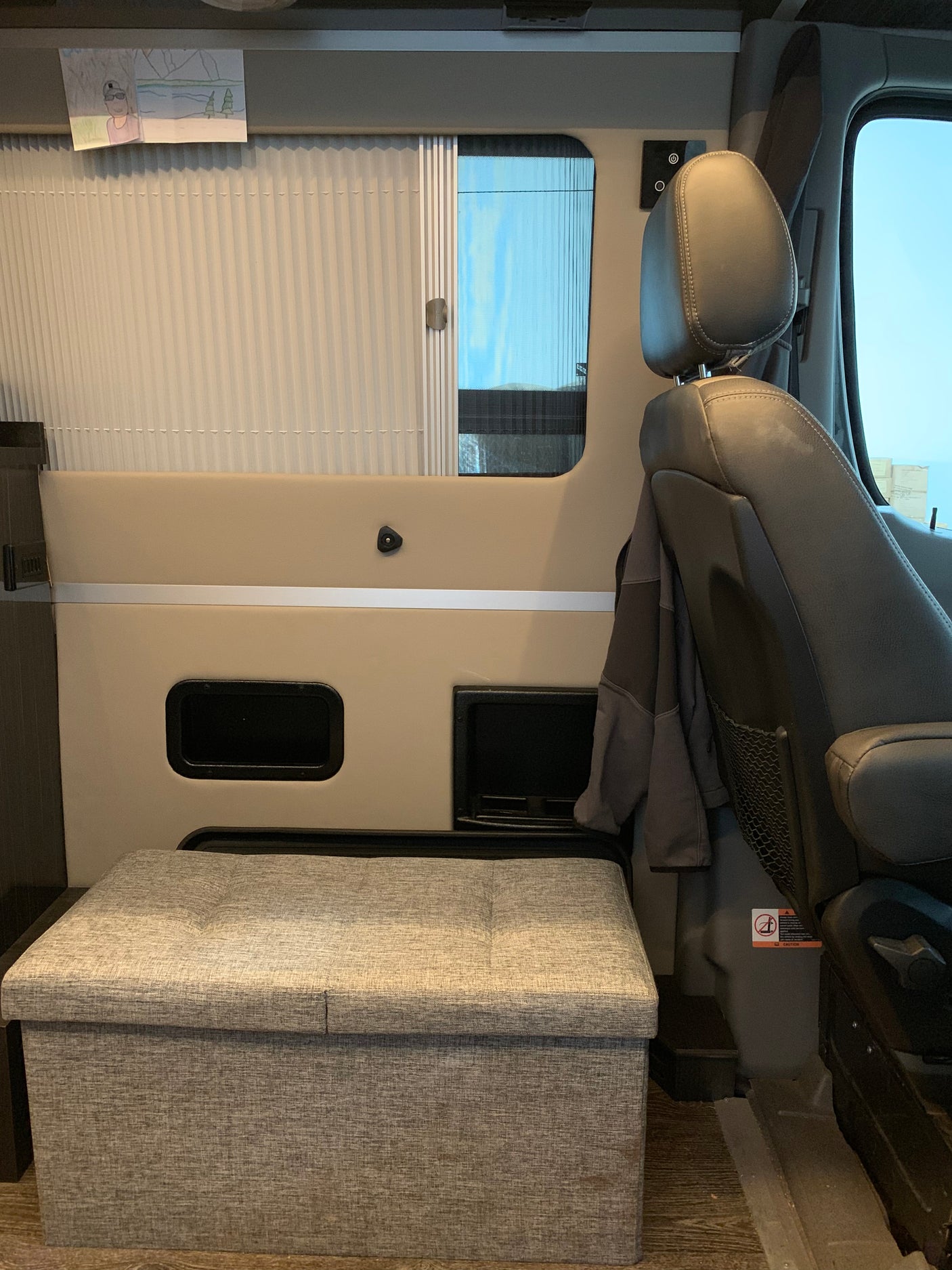 RV rental interior featuring a comfortable passenger seat, window with blind, and handy fabric storage ottoman.