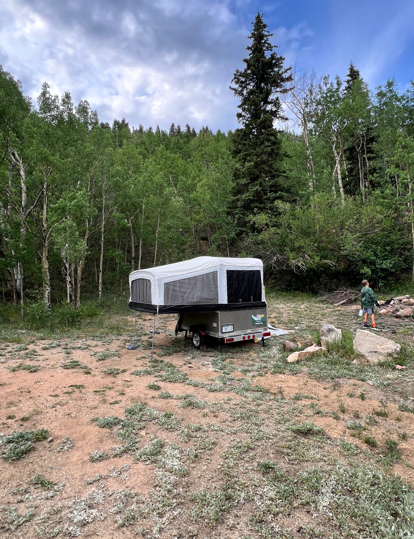 Pop-up camper RV rental set up in a wooded campsite with a child walking near a fire pit, surrounded by trees.