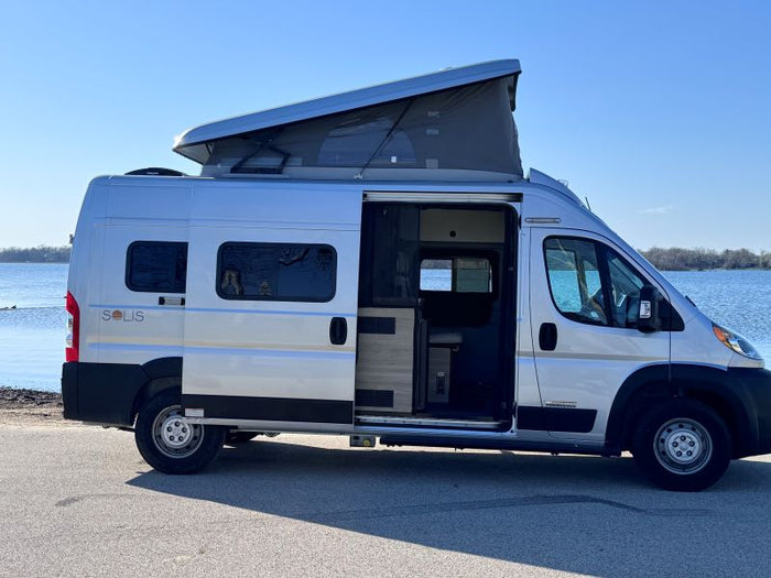 backed by a sunny forest. Ready for adventure.|Silver Solis pop-top camper van by a lake with its side door open