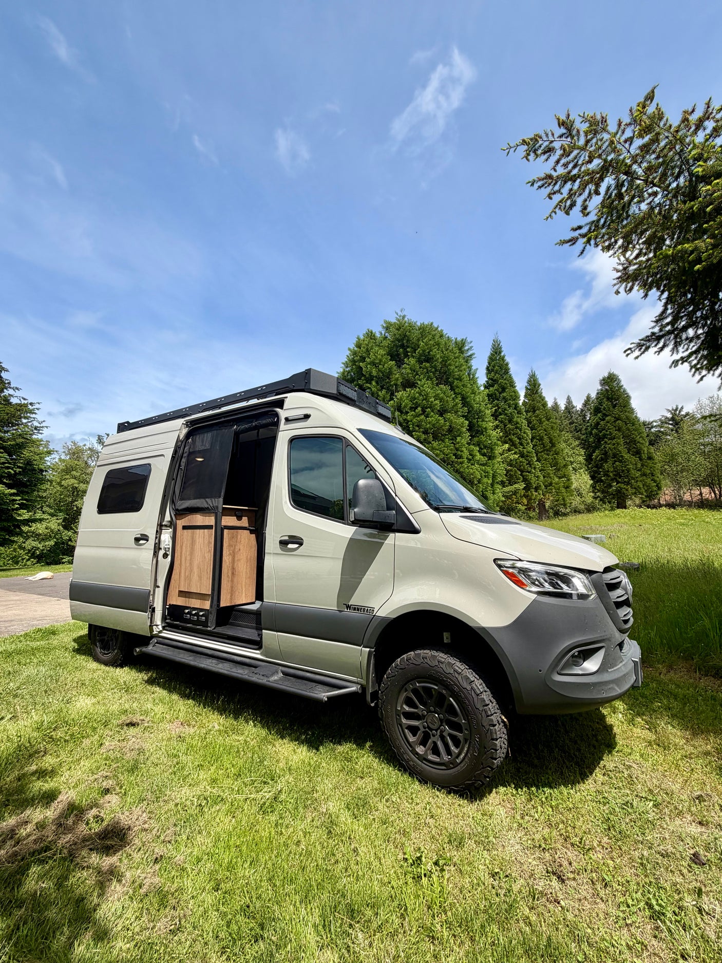 This tan Winnebago Revel camper van, great for rv rental, is parked on grass with its side door open, revealing a wood interior.