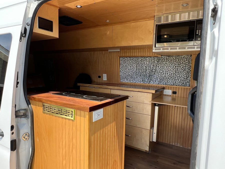 **RV rental.** This camper van kitchen features warm wooden finishes, microwave, cooktop, and storage solutions.