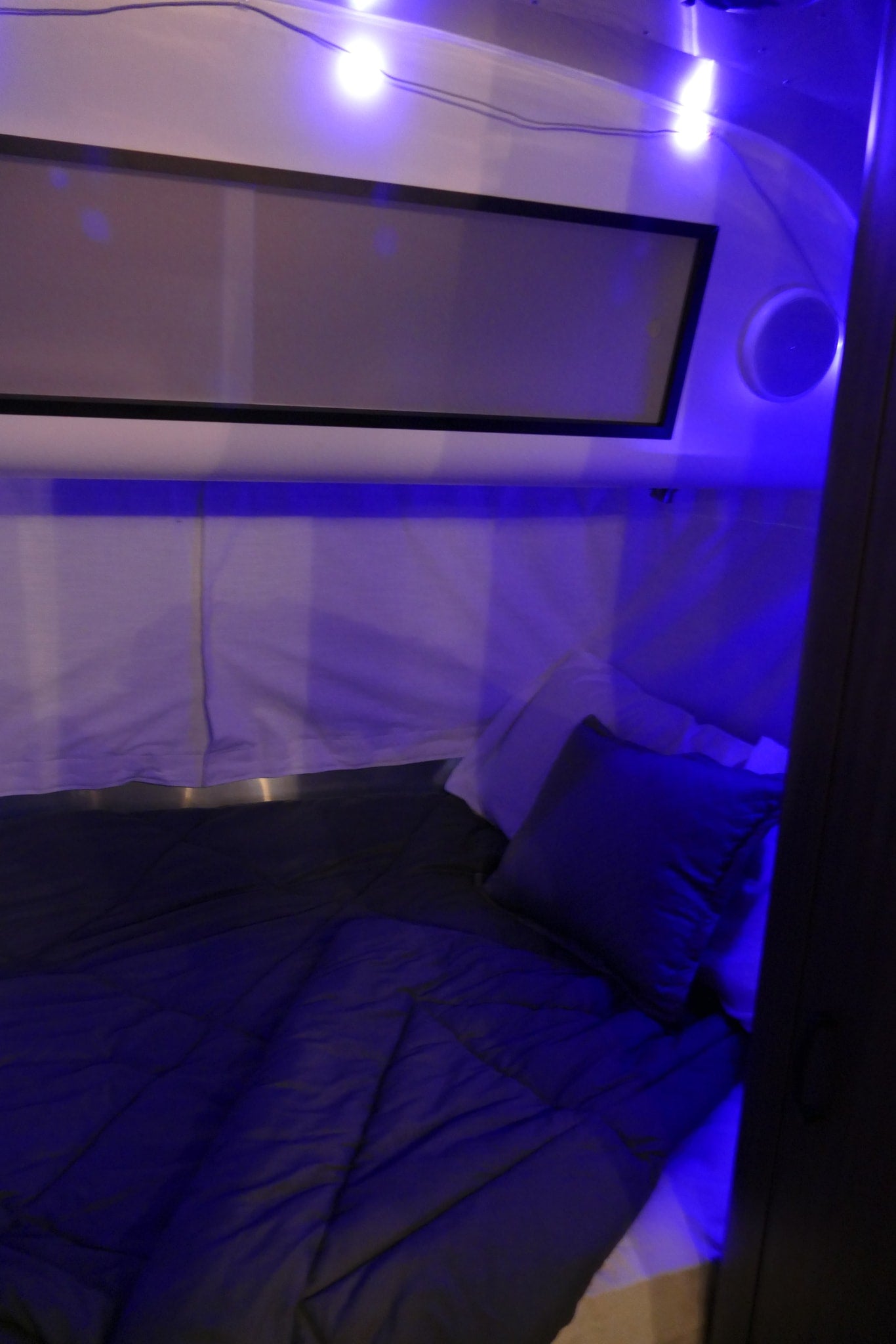 Inside an RV rental: a dimly lit bedroom with blue ambient lights, a made bed, pillows, and a window. Ready for your trip.