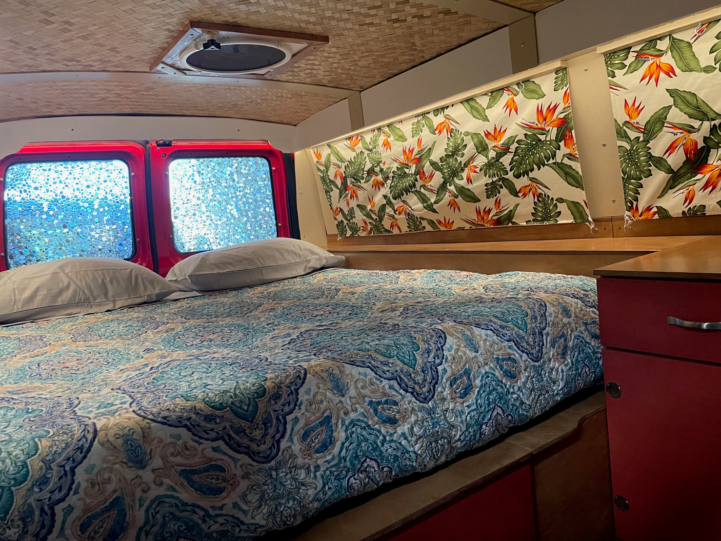 Inside a cozy RV rental: comfy bed with vibrant quilt, red window frames with textured glass, tropical curtains & bamboo ceiling.