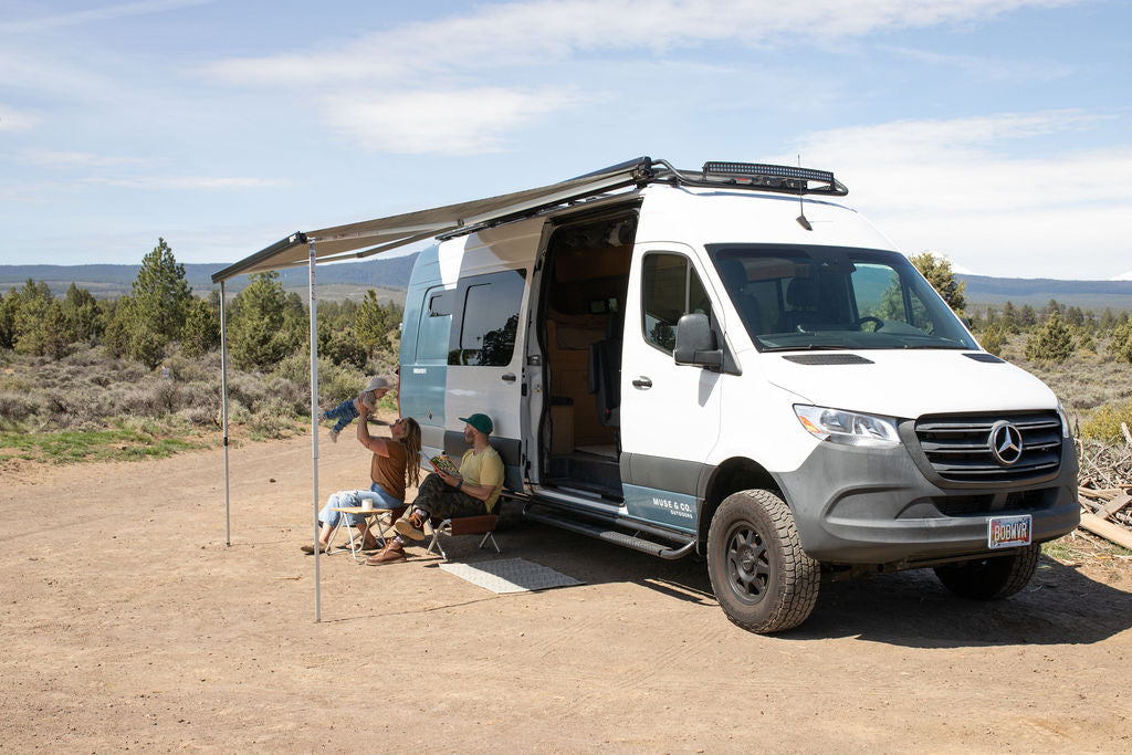 A family with a baby enjoys a relaxing day outside their white Mercedes Sprinter RV rental, parked with an awning extended in nature.