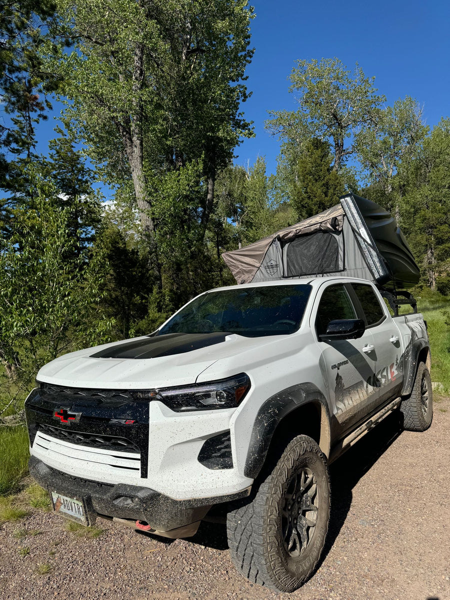 White Chevy Colorado ZR2 with rooftop tent set for an off-road adventure. Consider this flexible RV rental alternative.|Person smiles from white overland truck on rocky trail with rooftop tent. Your ultimate off-road rv rental adventure.|White pickup truck with rooftop tent & ladder