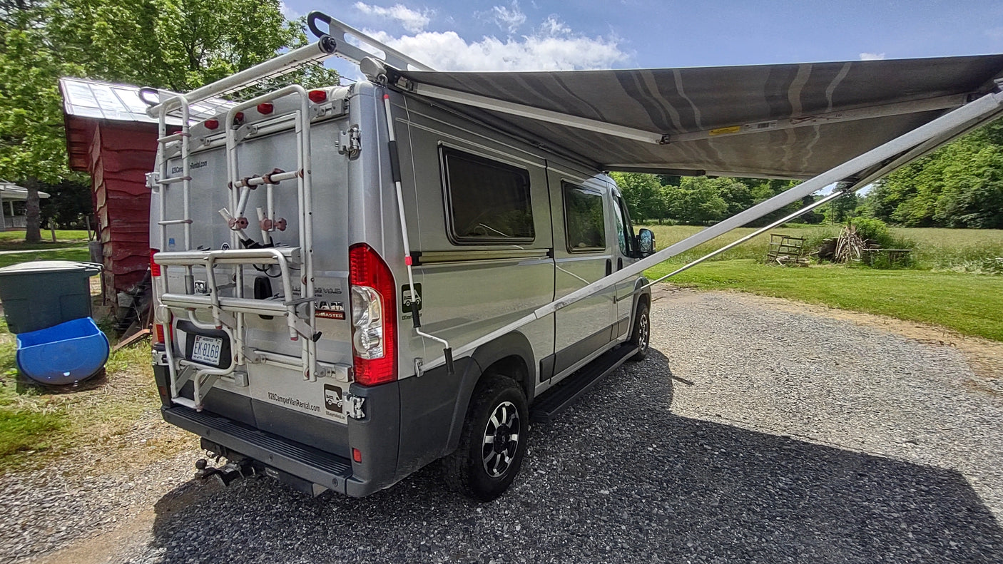 Parked silver RV rental camper van with a large awning extended, on a gravel drive with green fields.