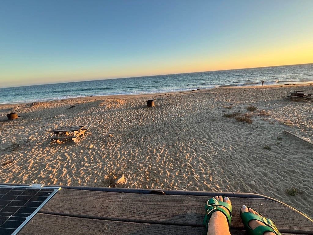 Feet up on our RV rental deck, enjoying a serene beach sunset. Solar panel visible, ocean waves, and a distant person on the sand.