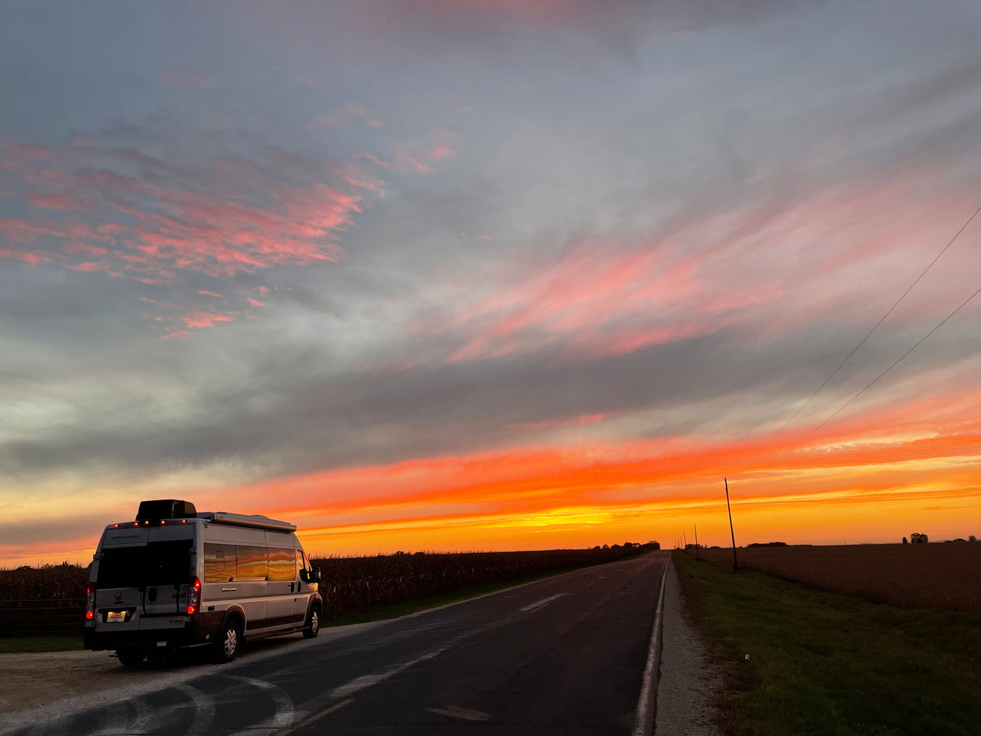 An RV rental is parked roadside at sunset, illuminating the sky with vibrant orange and pink clouds over vast fields and a long road.