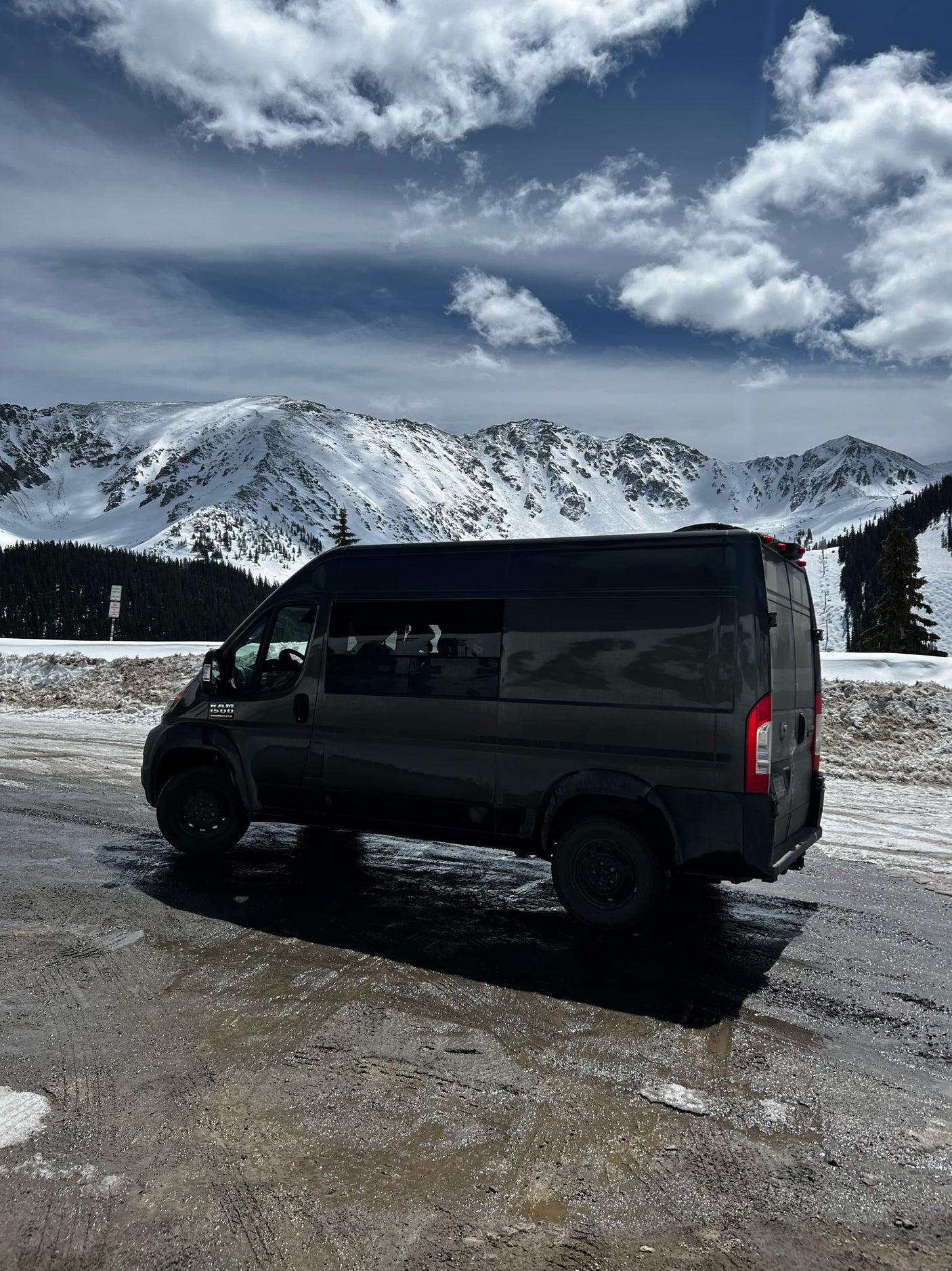 Explore winter peaks with this RV rental! Dark camper van parked on a snowy, muddy road under cloudy mountain skies.