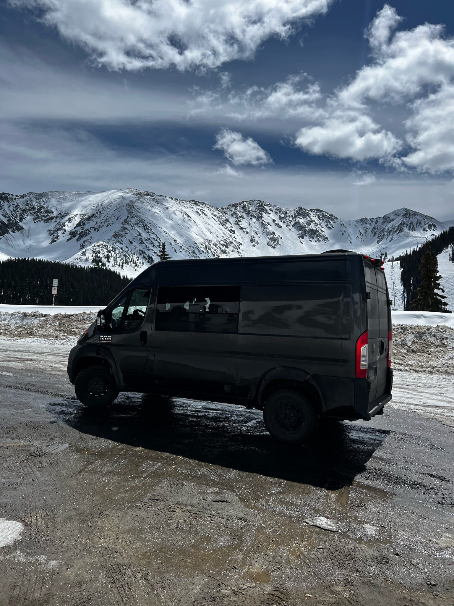 Explore winter peaks with this RV rental! Dark camper van parked on a snowy, muddy road under cloudy mountain skies.
