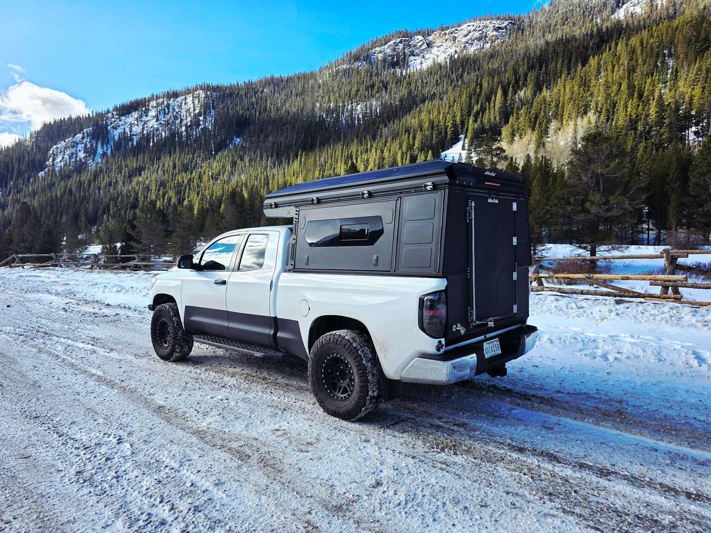White pickup truck with black overland camper on a snowy mountain road, surrounded by pine trees. Perfect for an rv rental adventure.