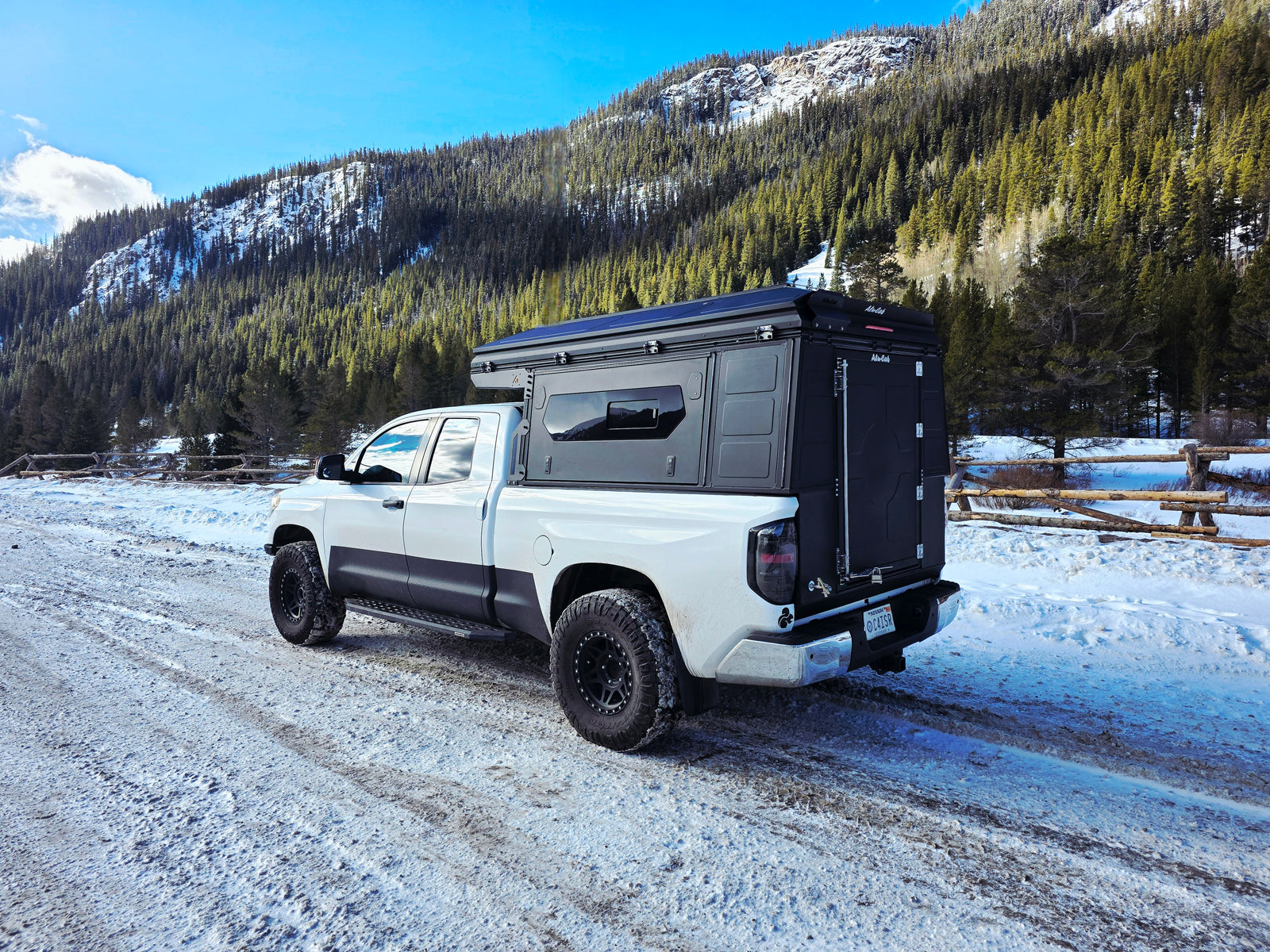 White pickup truck with black overland camper on a snowy mountain road, surrounded by pine trees. Perfect for an rv rental adventure.