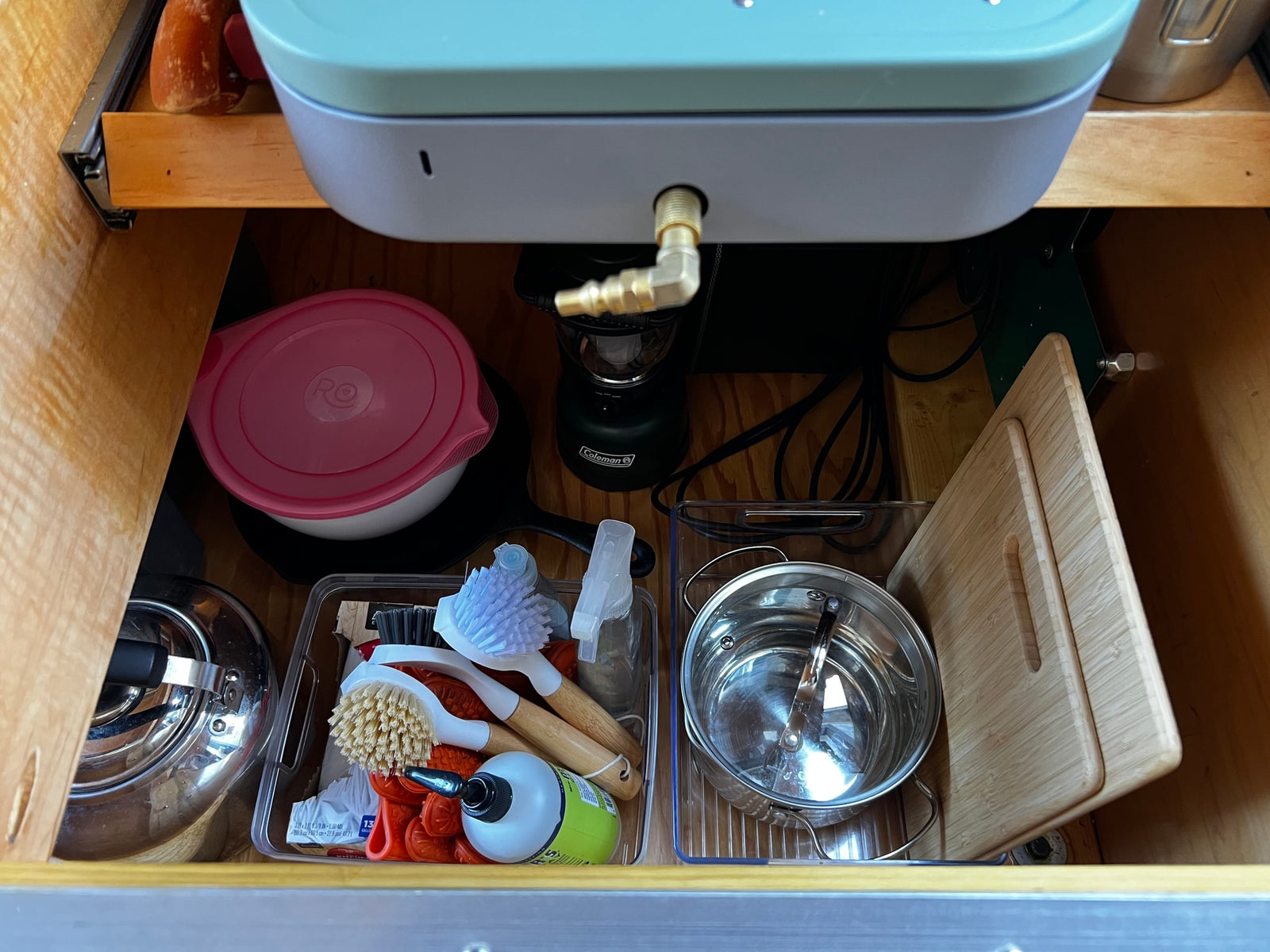 RV rental storage: An organized cabinet with cookware (pot, pan, cutting boards), cleaning supplies (brushes, soap), and a Coleman lantern.