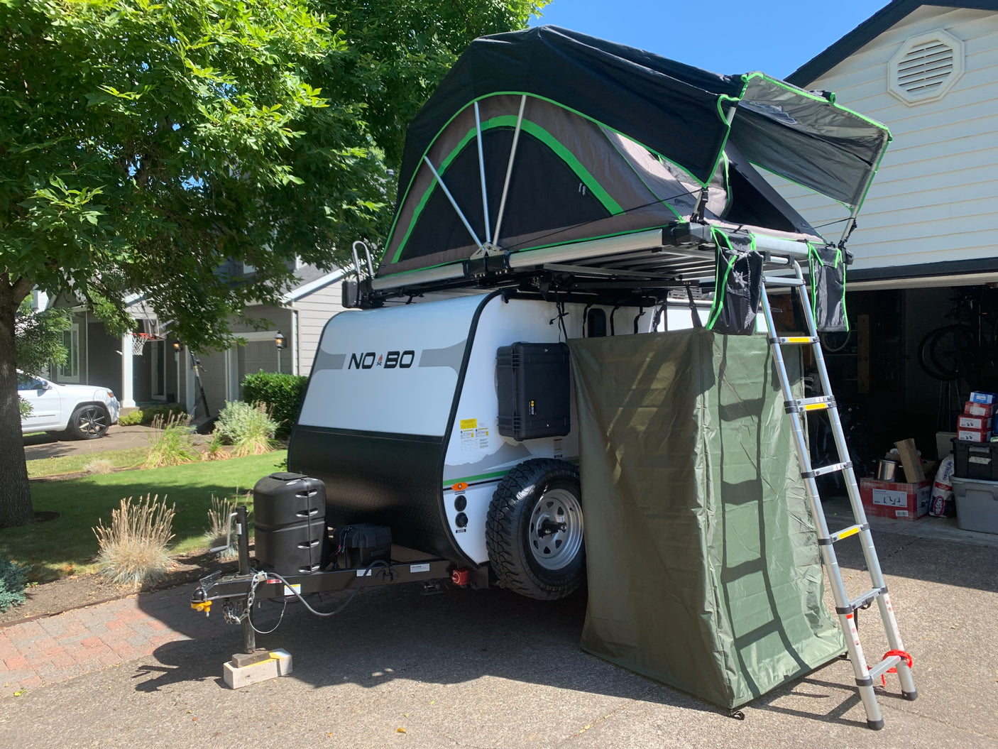 Adventure-ready rv rental: A white NO BO travel trailer with a rooftop tent and privacy annex is set up in a driveway, ideal for outdoor travel.