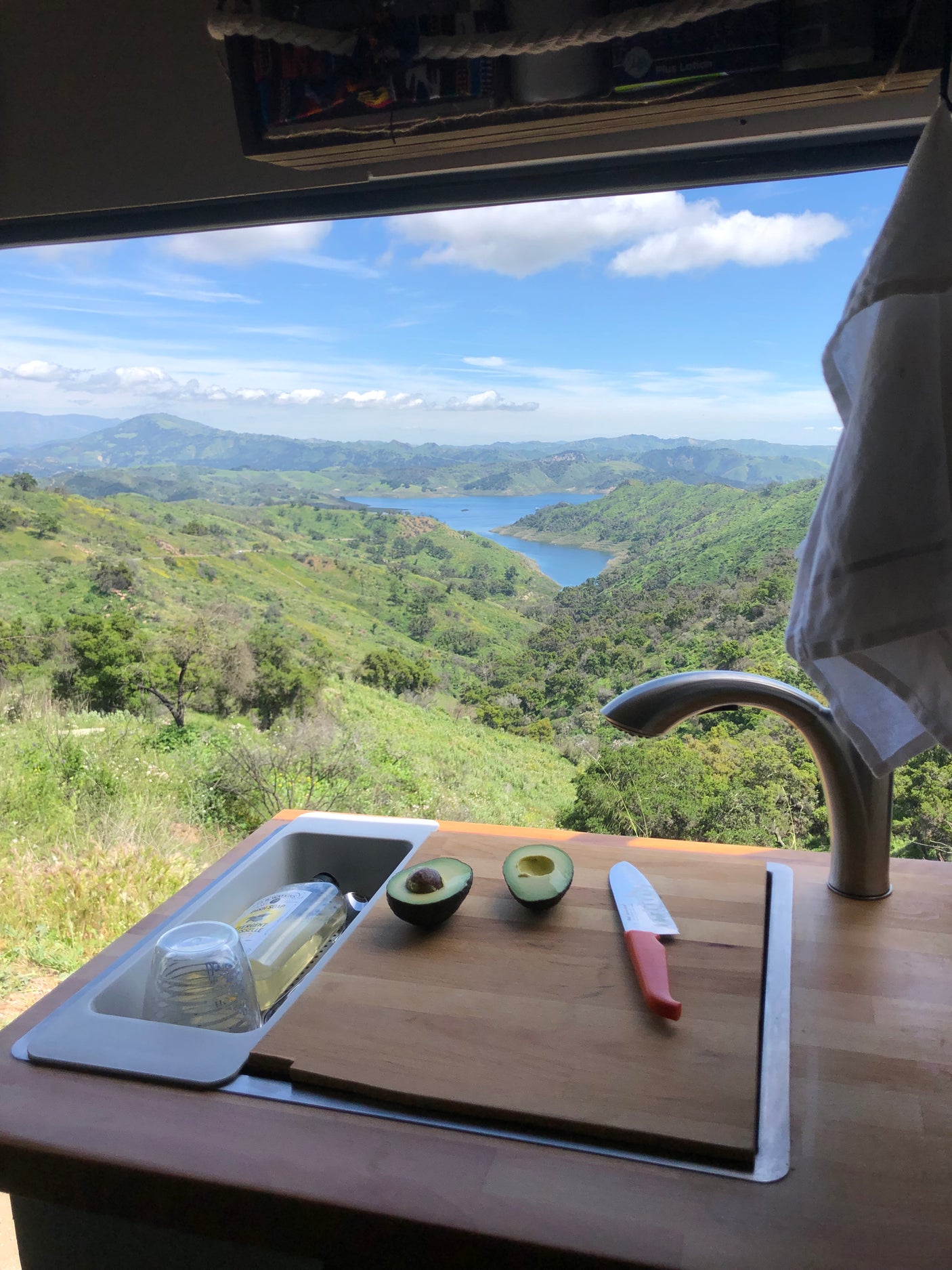 RV rental kitchen: preparing avocado with a stunning view of green hills and a serene blue lake.