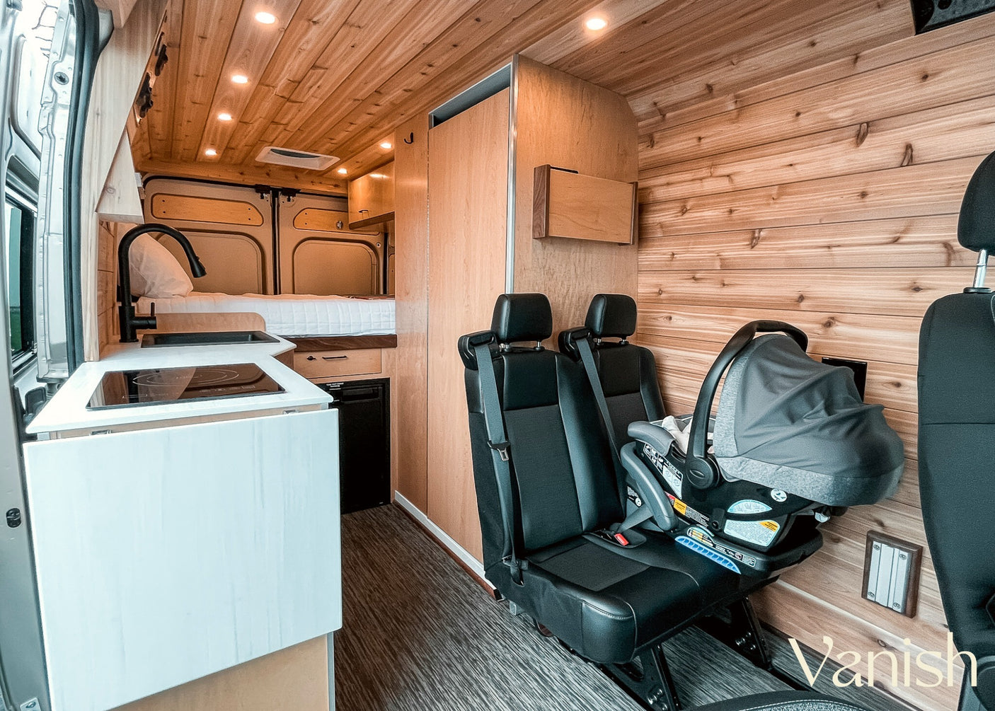 This RV rental interior features a cozy kitchen, bed, passenger seats, and a baby car seat, perfect for family vanlife.