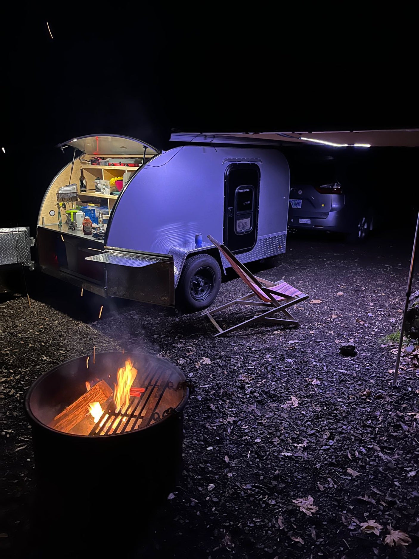 An illuminated teardrop rv rental camping setup at night, with an open kitchen, fire pit, and extended awning.