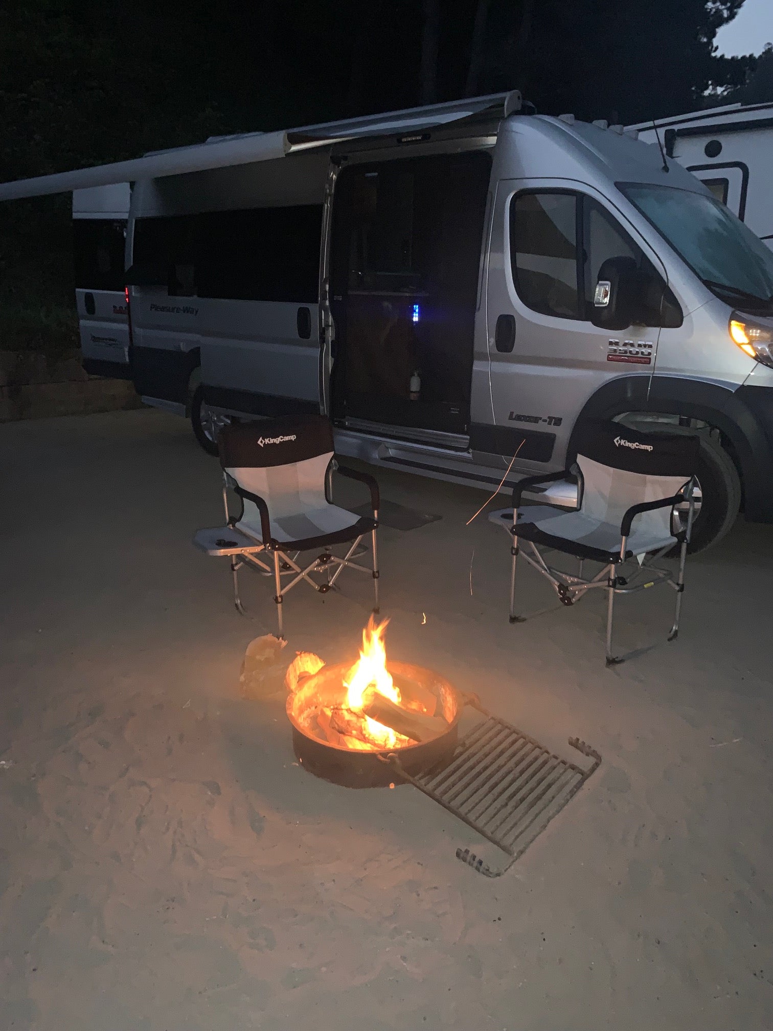 Cozy RV rental campsite at night: a silver campervan, two chairs, and a warm, glowing campfire on sandy ground.