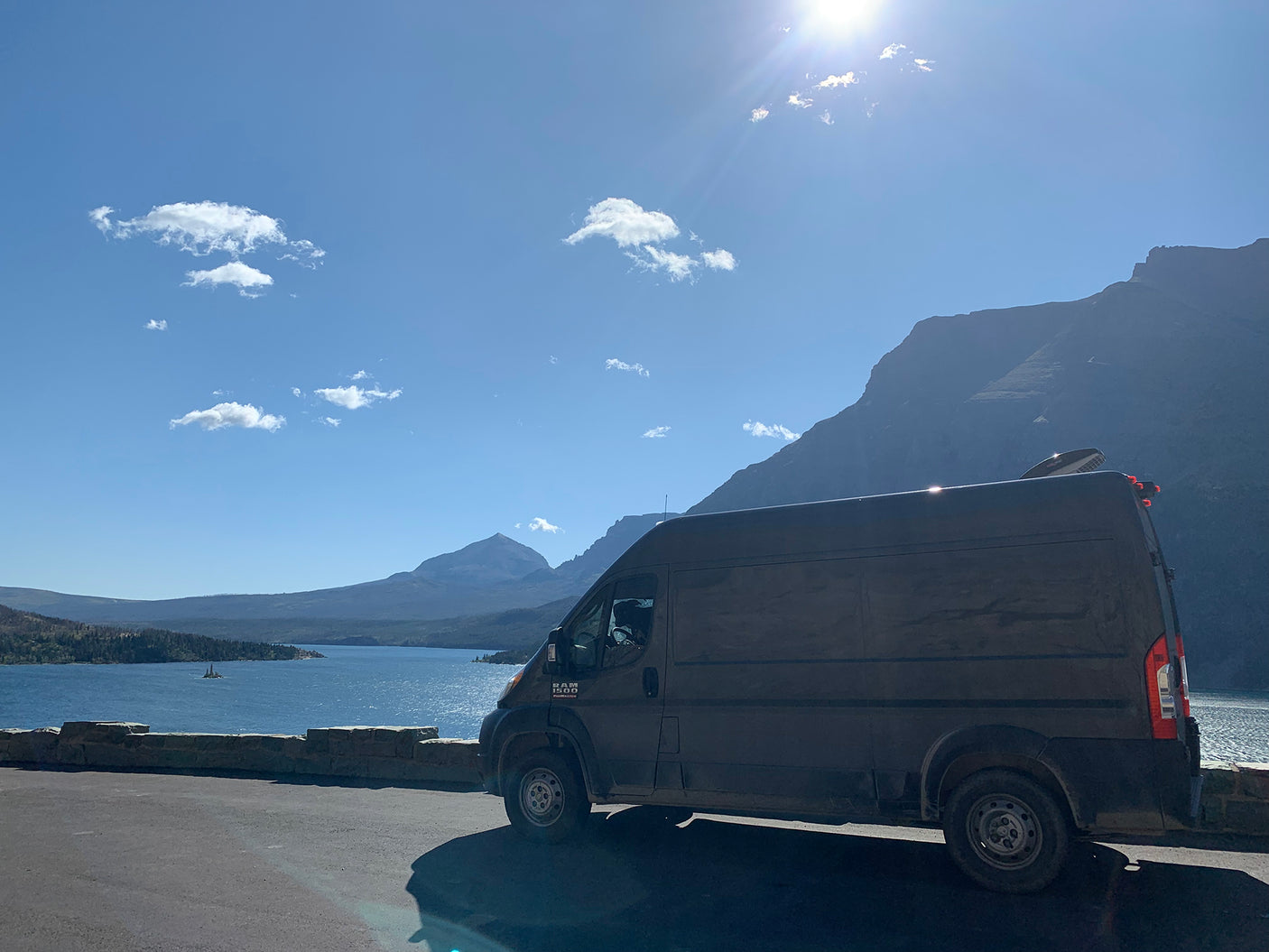 Dark RV rental van parked beside a stone wall, overlooking a vast blue lake & mountains under a bright sky.