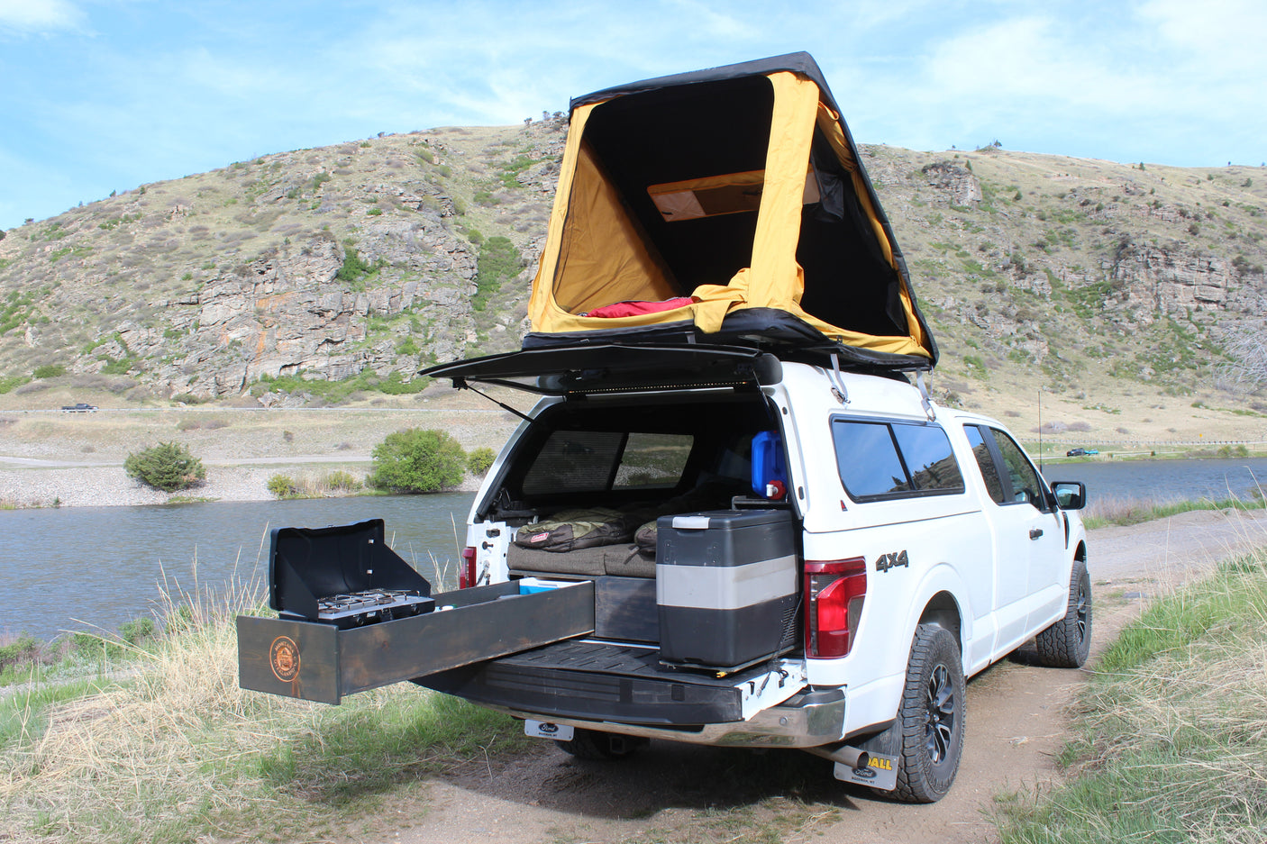 Overlanding truck with rooftop tent & outdoor kitchen by a scenic lake, perfect for an RV rental adventure.