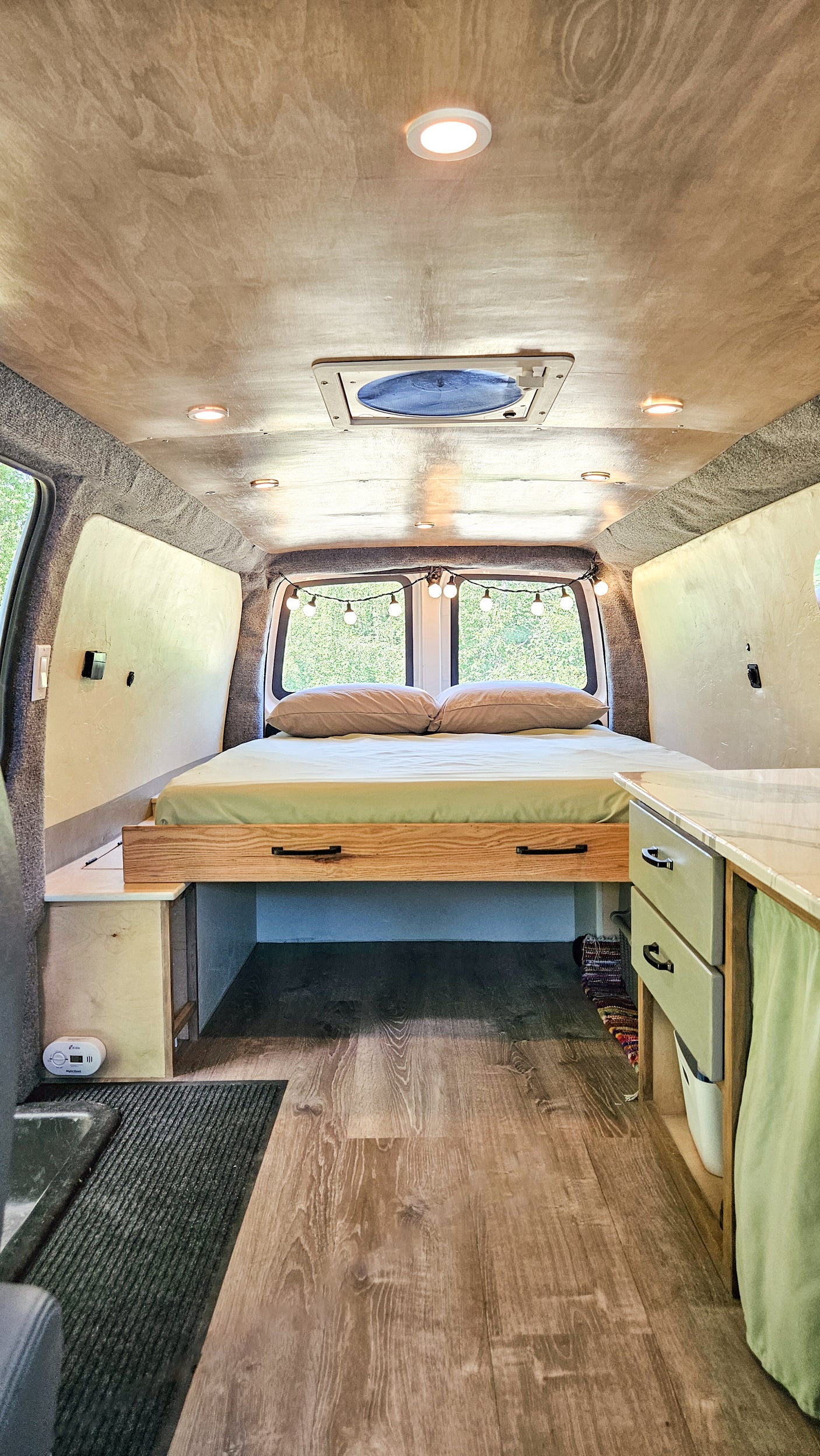 RV rental: Inside a cozy camper van featuring a rear bed, wood-look flooring, custom storage, and ceiling lighting.
