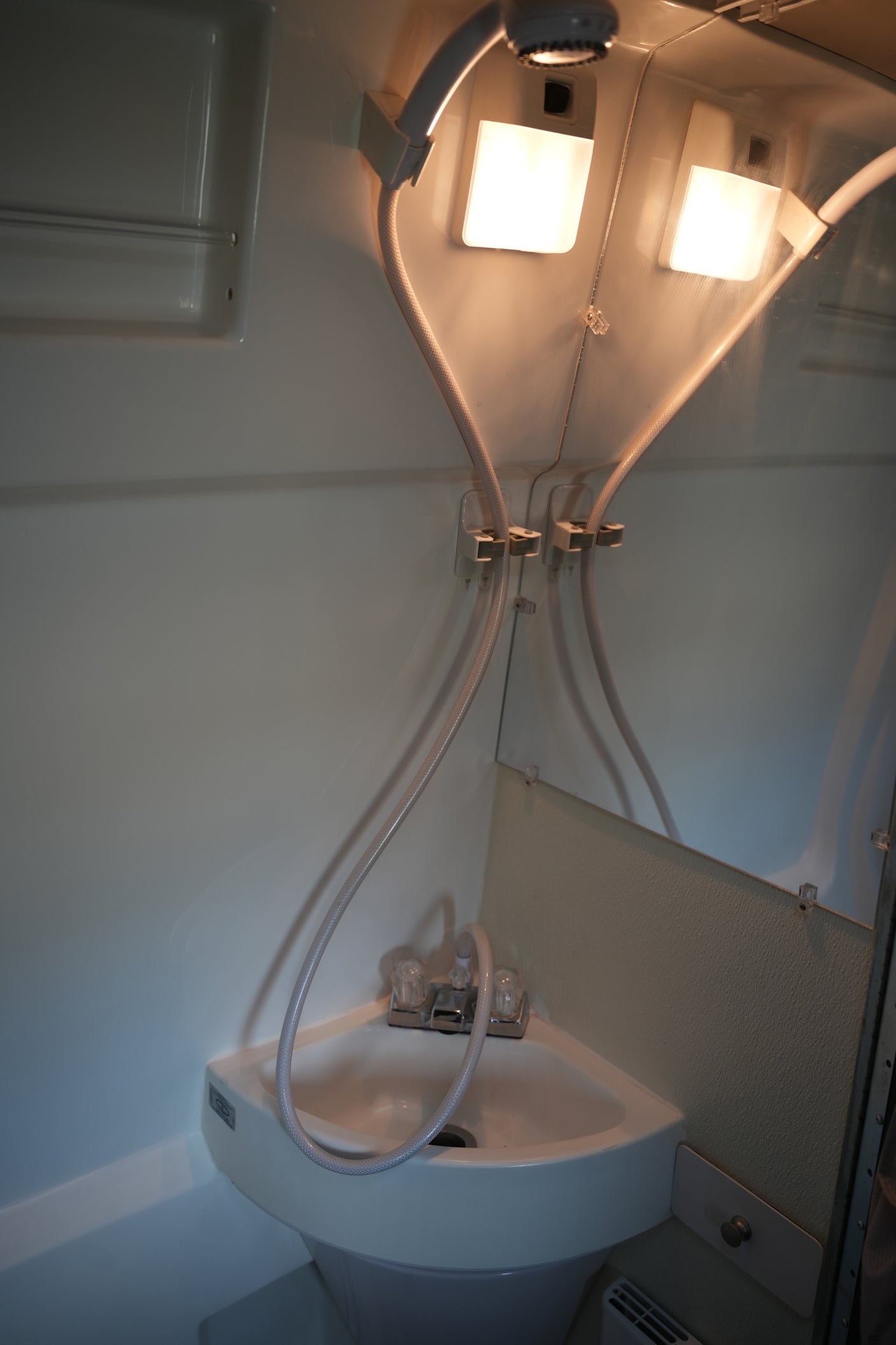 Inside an RV rental: a compact bathroom with a corner sink, dual-purpose shower head, mirror, and two bright overhead lights.
