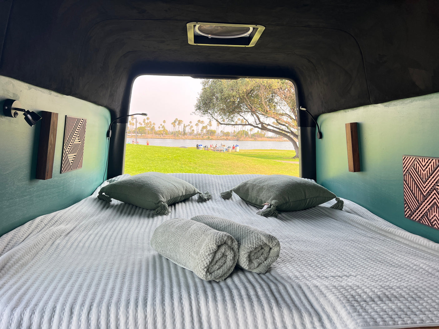 Inside a cozy RV rental: a neatly made bed overlooks a beautiful lake, green park, and palm trees under a bright sky.