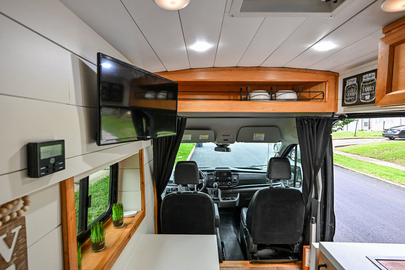View into an RV rental van's interior: living space with a wall-mounted TV, built-in storage, and the front cockpit.