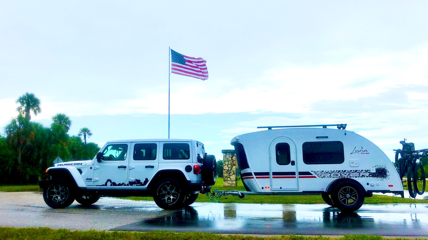 A white Jeep Wrangler tows a sleek Luna Rover RV rental on wet pavement, set against an American flag. Perfect for road trips.
