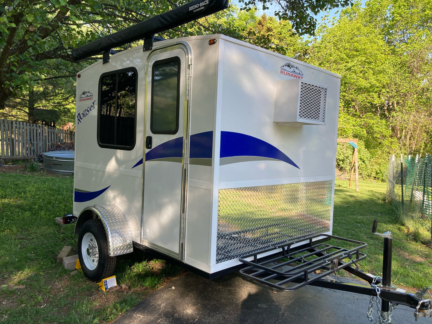A white compact Runaway Rouser RV rental teardrop camper, featuring an awning, AC, and front cargo rack, parked on asphalt.