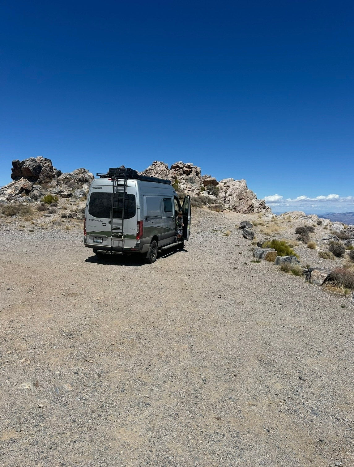 Grey RV rental camper van parked on a gravel path in a rocky desert, side door open under a vast blue sky. Adventure travel.