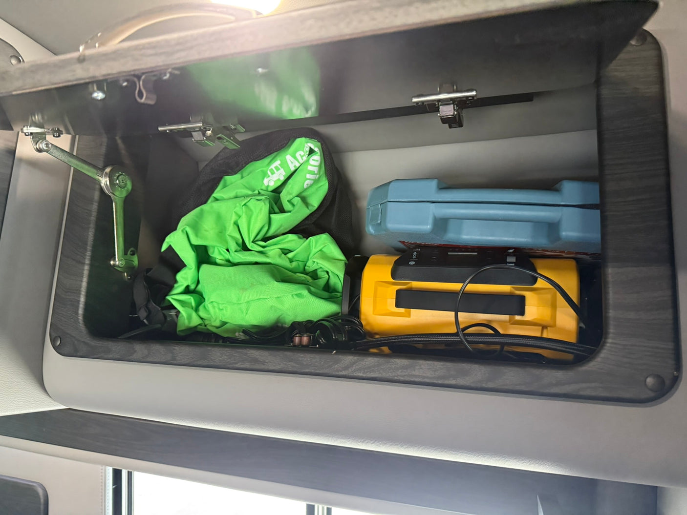 Overhead cabinet in an RV rental, holding a green emergency bag, blue case, and a yellow portable air compressor.