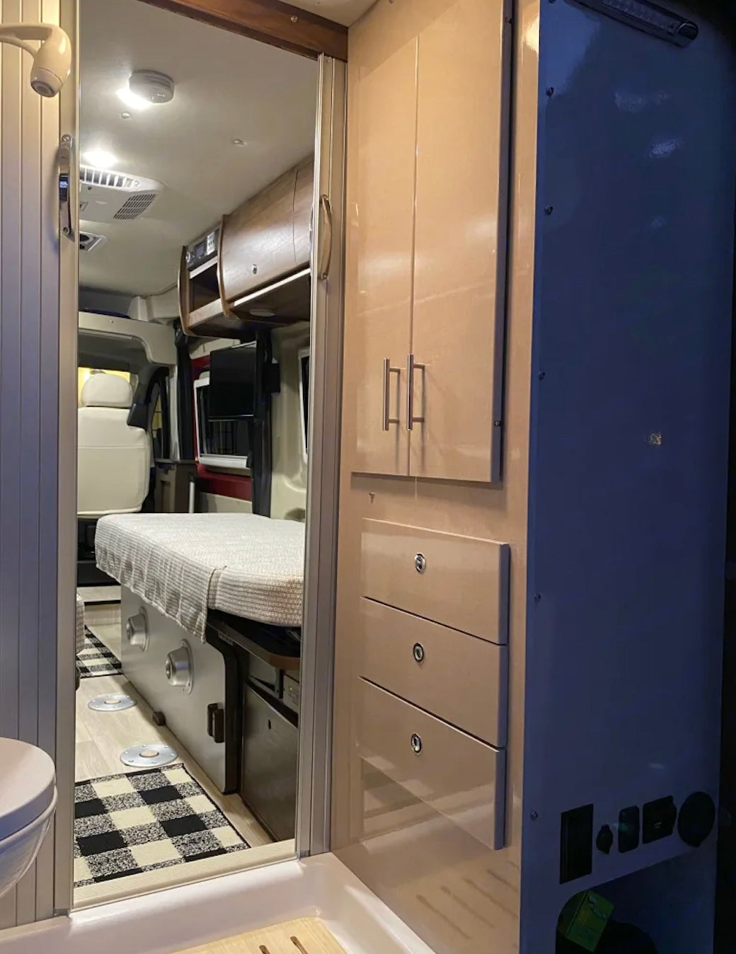 Explore this rv rental's compact interior: shower/toilet, sleek storage, and bed with checkered rug in the main living space.