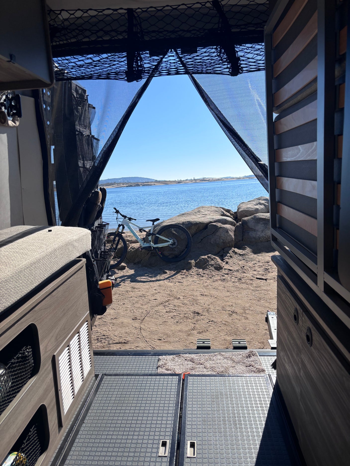 Inside an RV rental: Looking out at a mountain bike on sandy shore by a lake under clear blue sky.