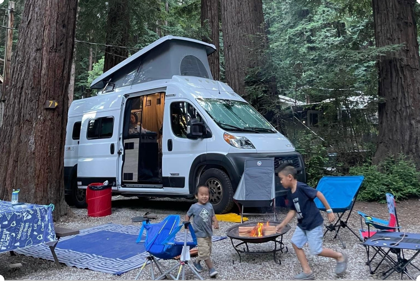Family fun at a redwood campsite with their rv rental. Kids play by the fire, campervan door open, enjoying nature.