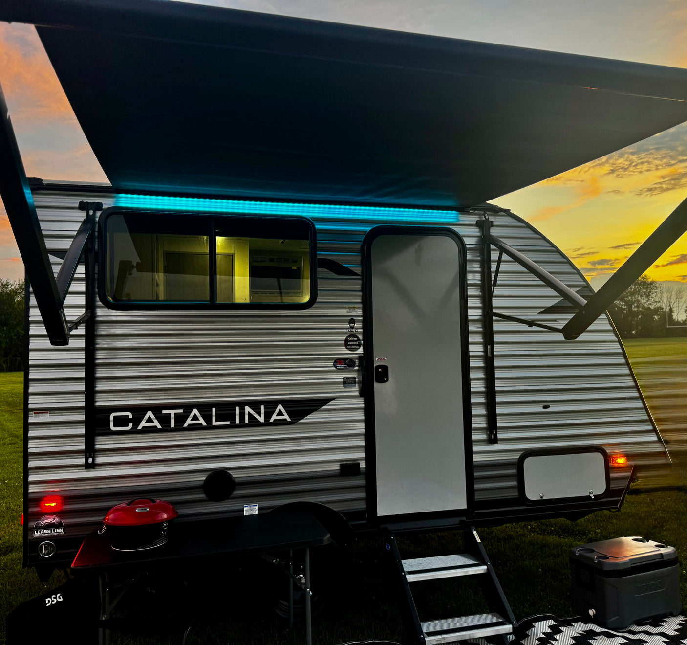 Evening campsite view: a Catalina RV rental with blue awning lights, grill, and cooler under a vibrant sunset.