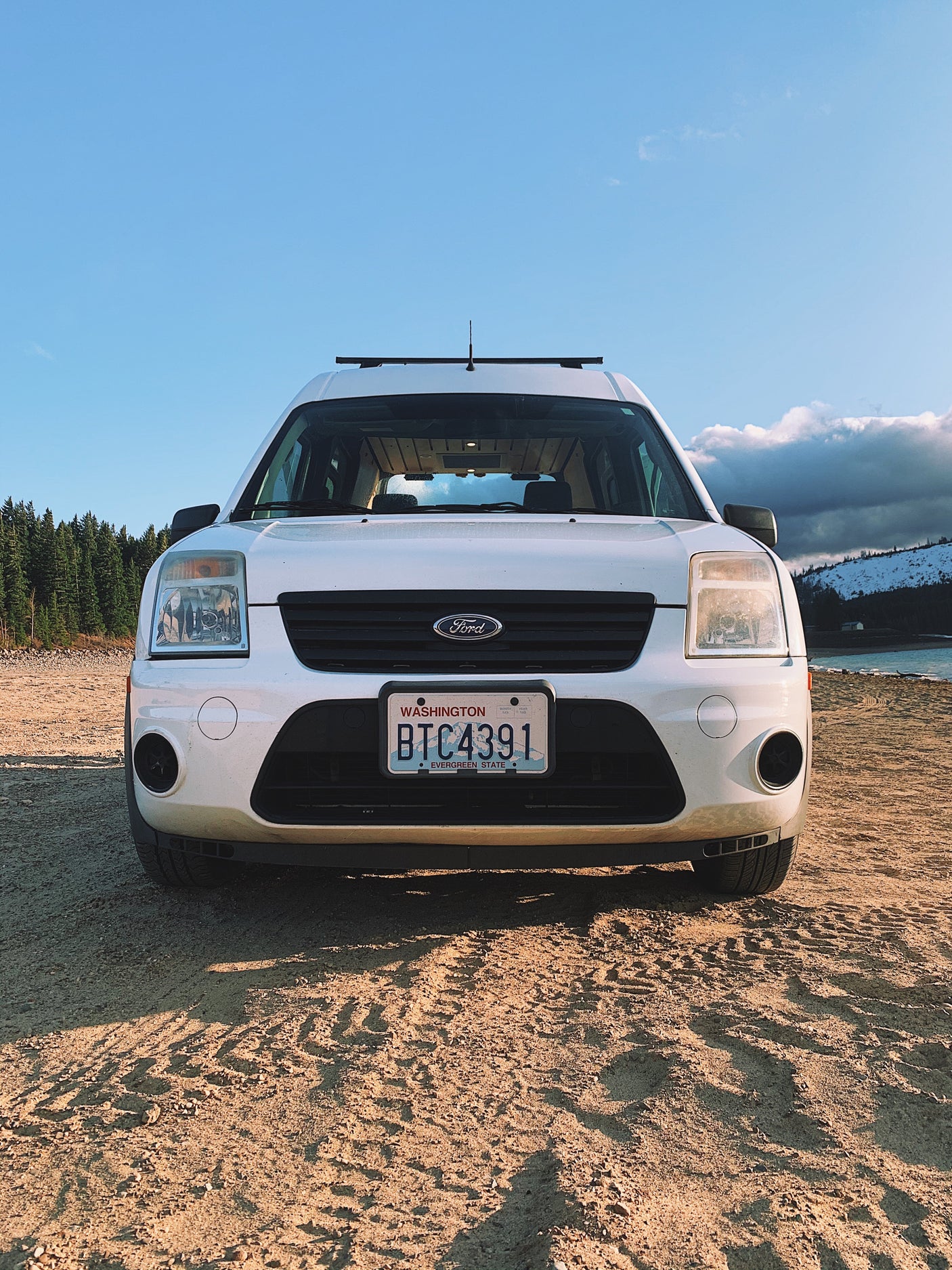 White Ford Transit Connect RV rental on a dirt road by a lake and forest. Ideal for camping and road trip adventures.