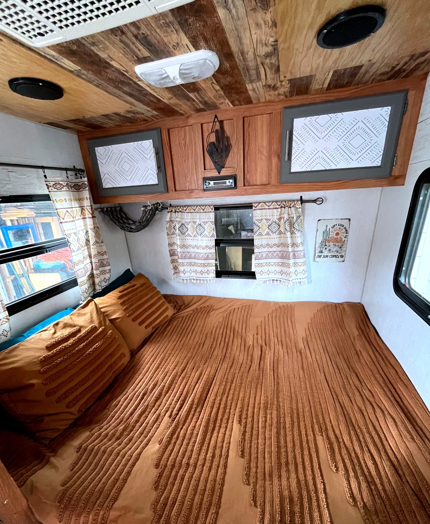 This cozy RV rental interior features a full bed with textured covers, patterned curtains, and custom wooden ceilings.