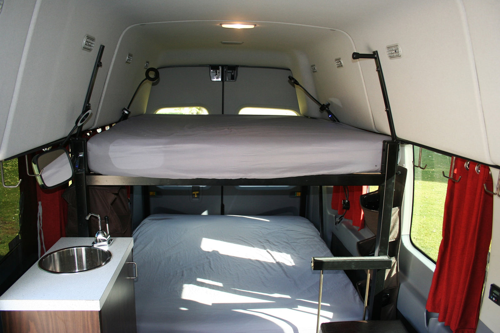 RV rental: Camper van interior with stacked bunk beds and a small sink on the left, red curtains on the right window.