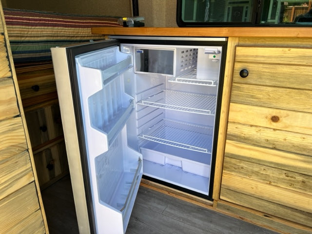 RV rental: Bright, open refrigerator with empty shelves in a custom wood RV interior, showing cabinets, drawers, and a bed.