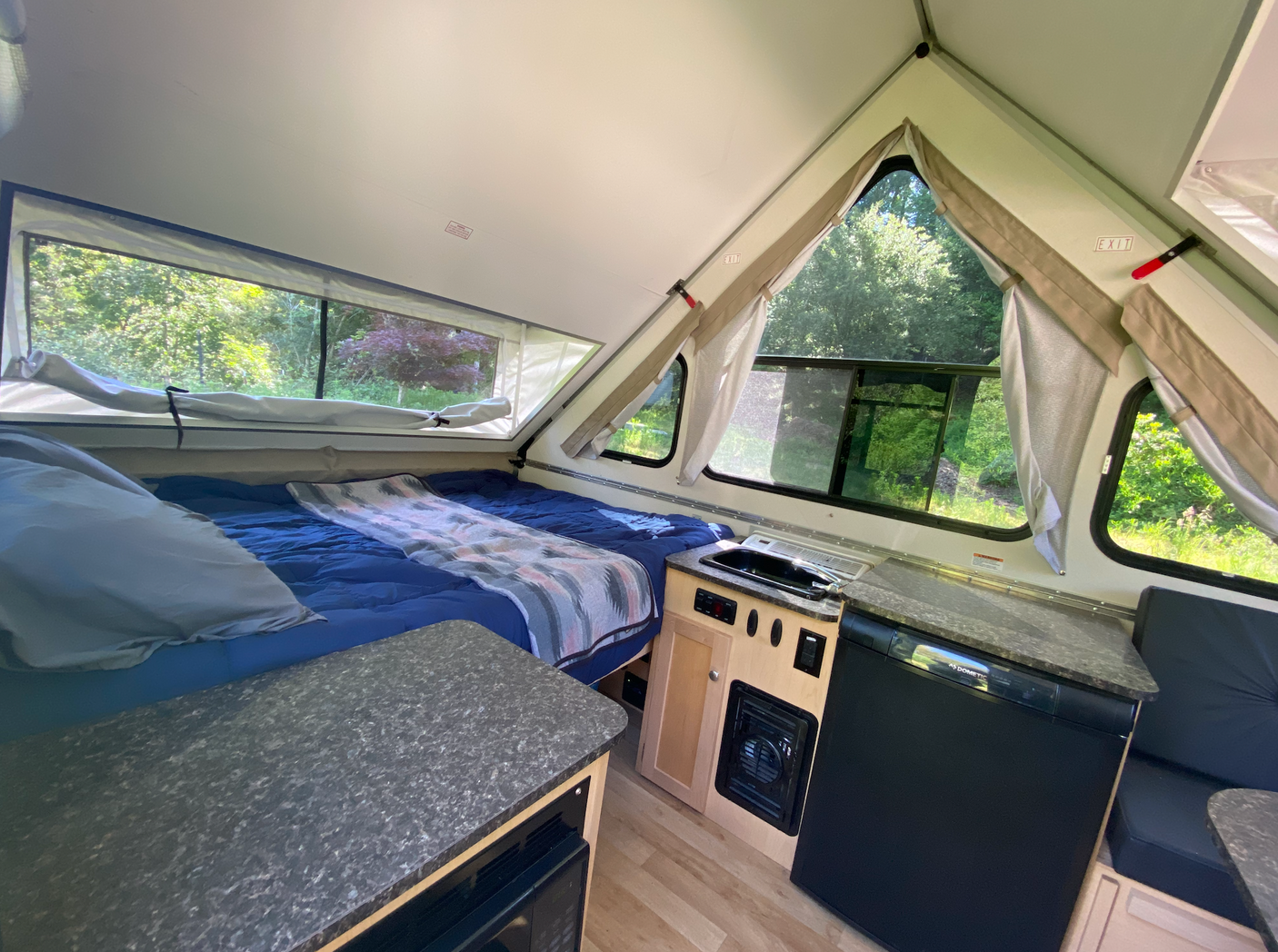 A-frame RV rental interior: A cozy bed, compact kitchenette with fridge & microwave, and bright windows overlook nature.