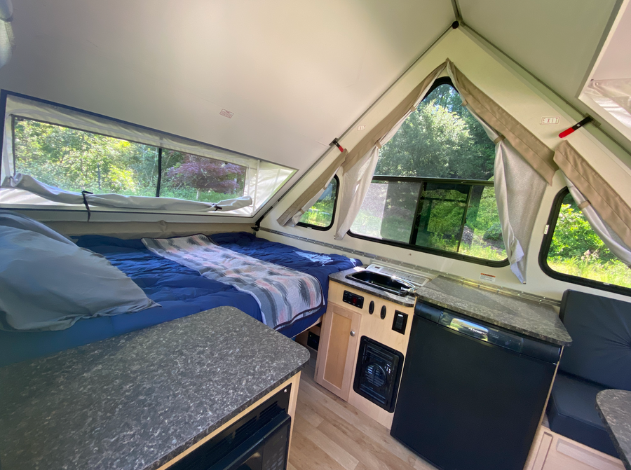 A-frame RV rental interior: A cozy bed, compact kitchenette with fridge & microwave, and bright windows overlook nature.