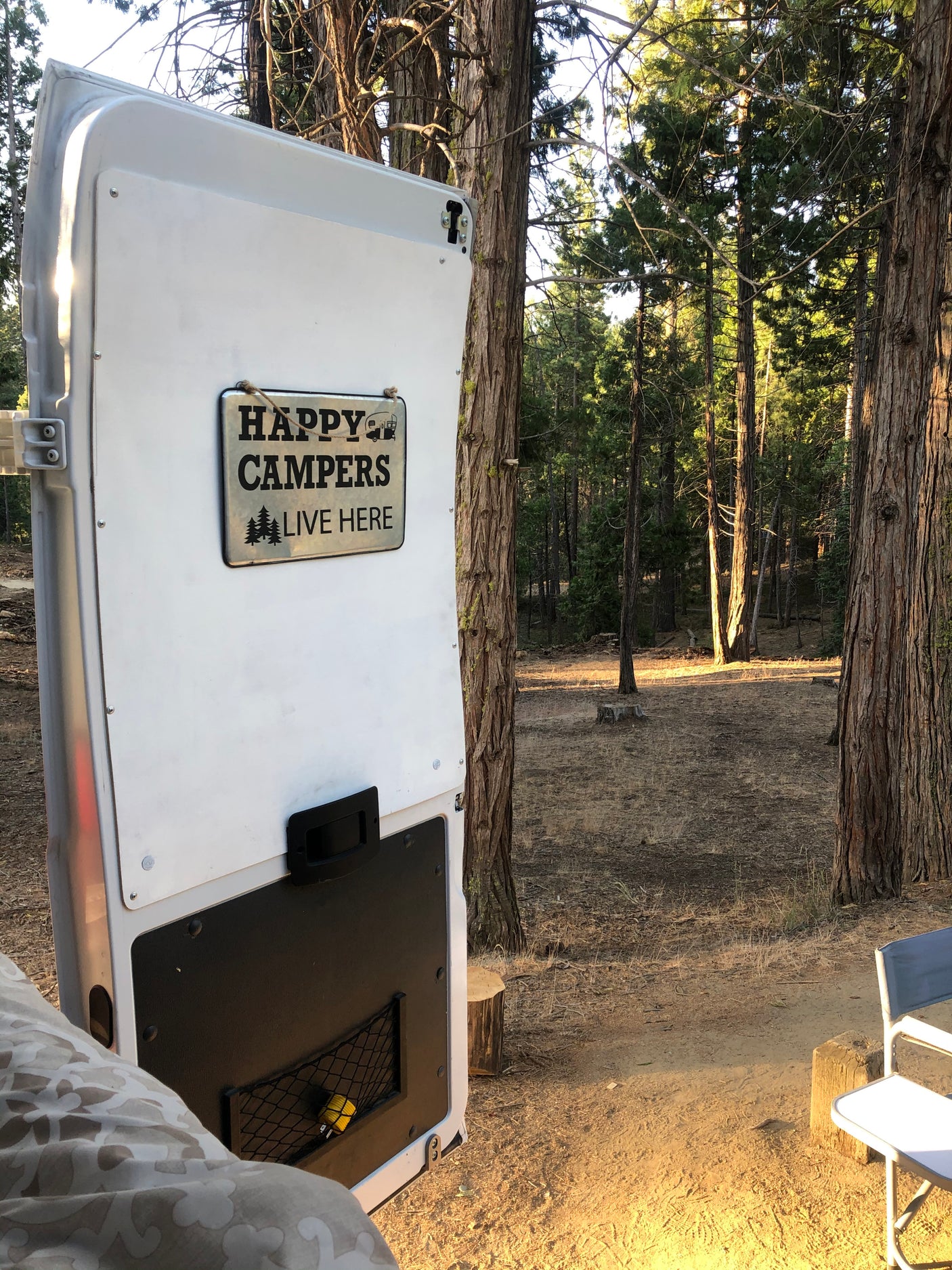 White RV rental door with a 'Happy Campers Live Here' sign, open to a peaceful forest setting with tall trees and dry ground.