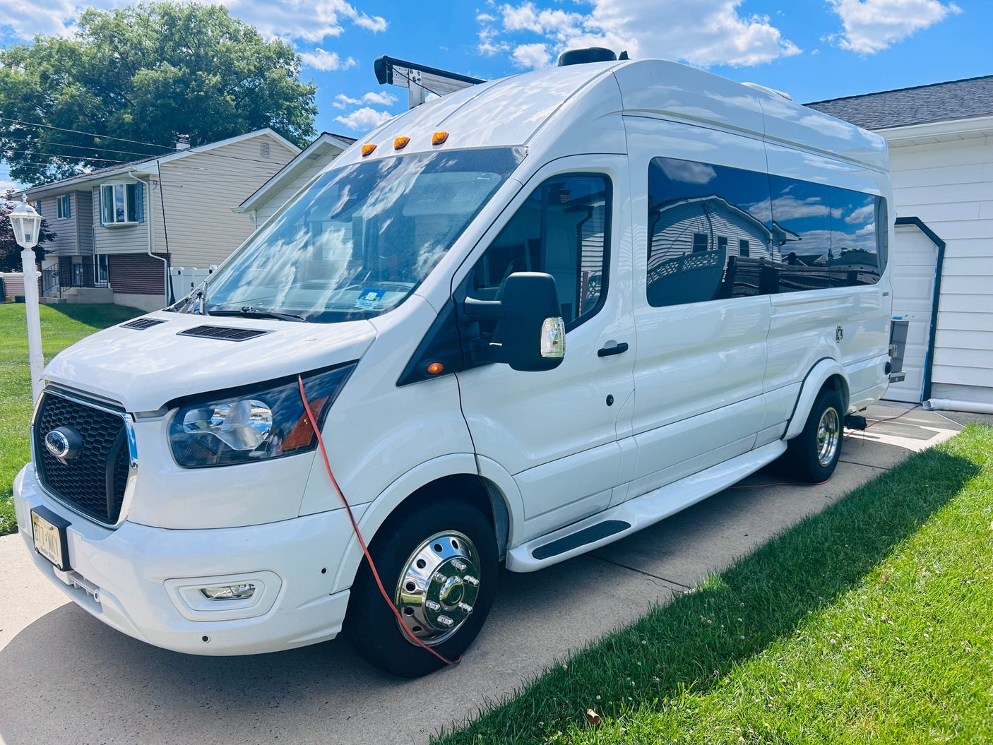 White Ford Transit camper van for rv rental, parked on a sunny driveway beside a house. Clouds reflect in windows.