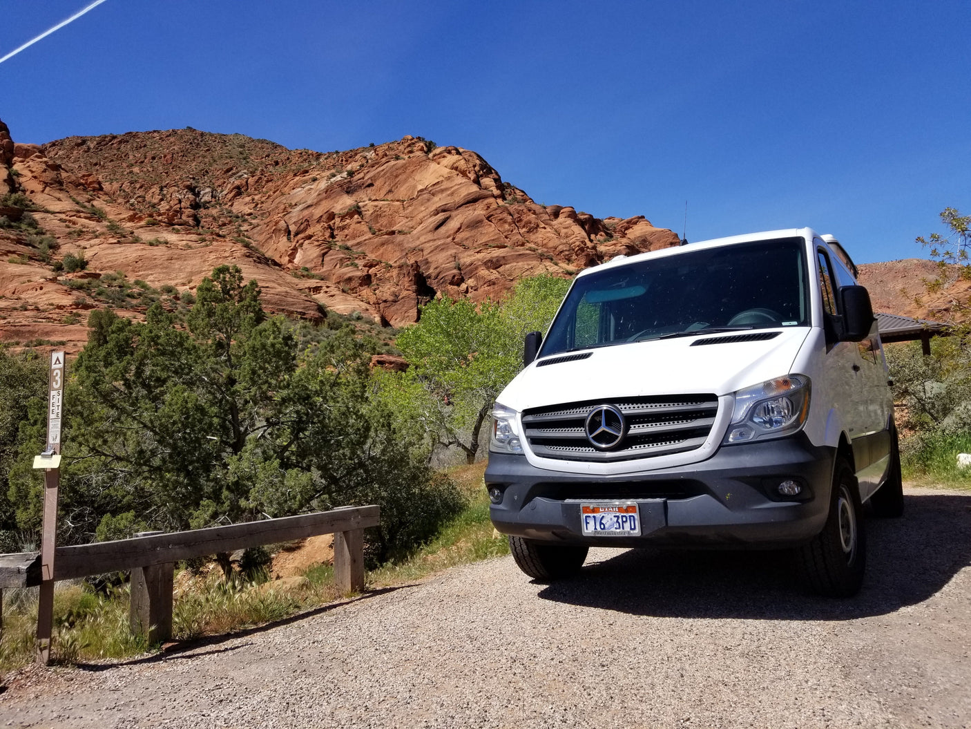 A white Mercedes-Benz Sprinter RV rental van parked in a scenic red rock campsite with green trees under a clear blue sky.
