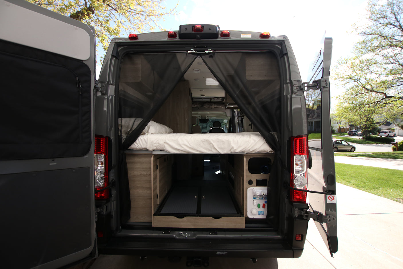 This rv rental's open back shows a cozy bed, storage, and utility panel, protected by a mosquito net, ideal for travel.