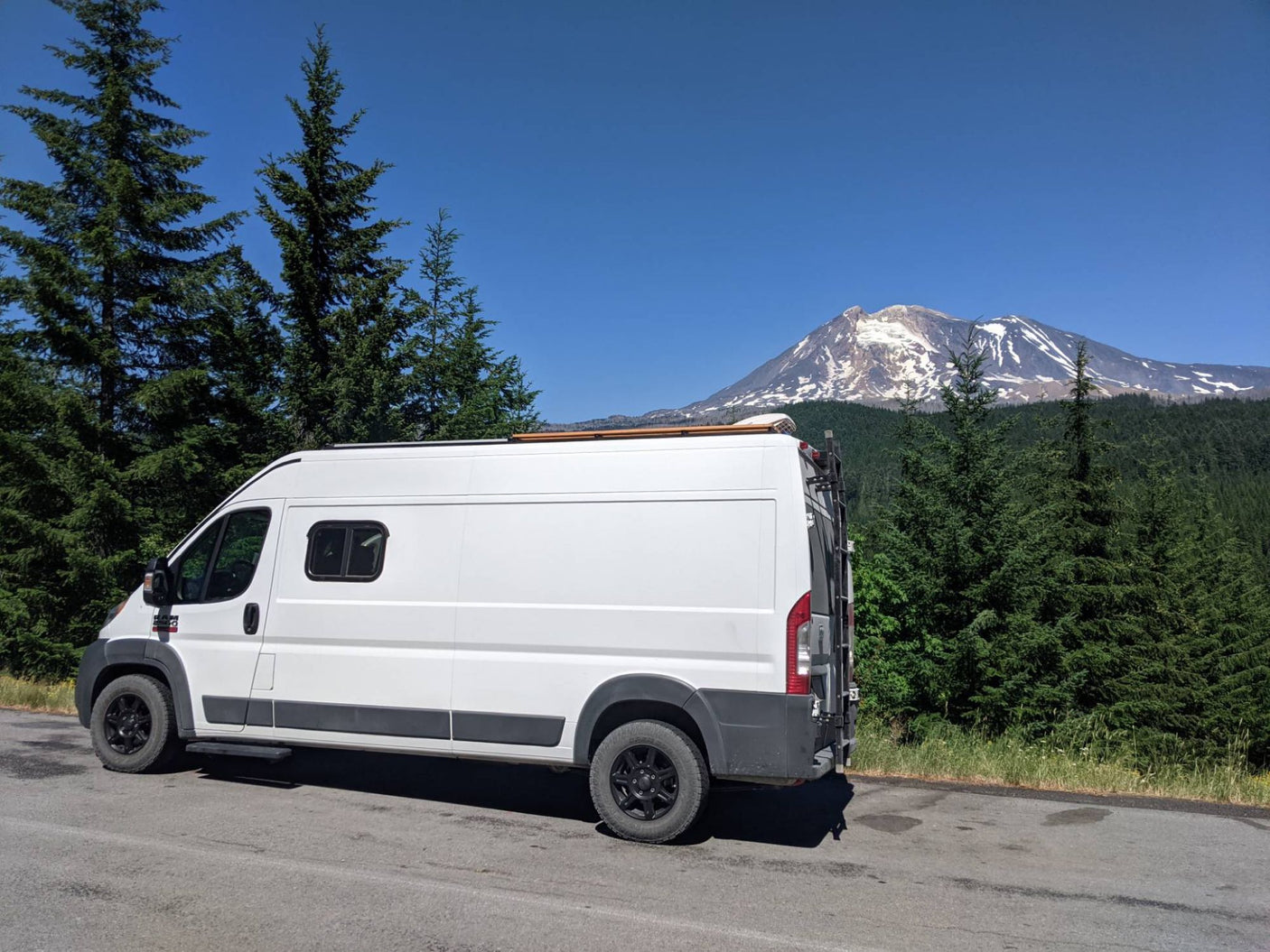 Discover the PNW! White RV rental parked roadside amidst tall pines, with a stunning snow-capped mountain backdrop.