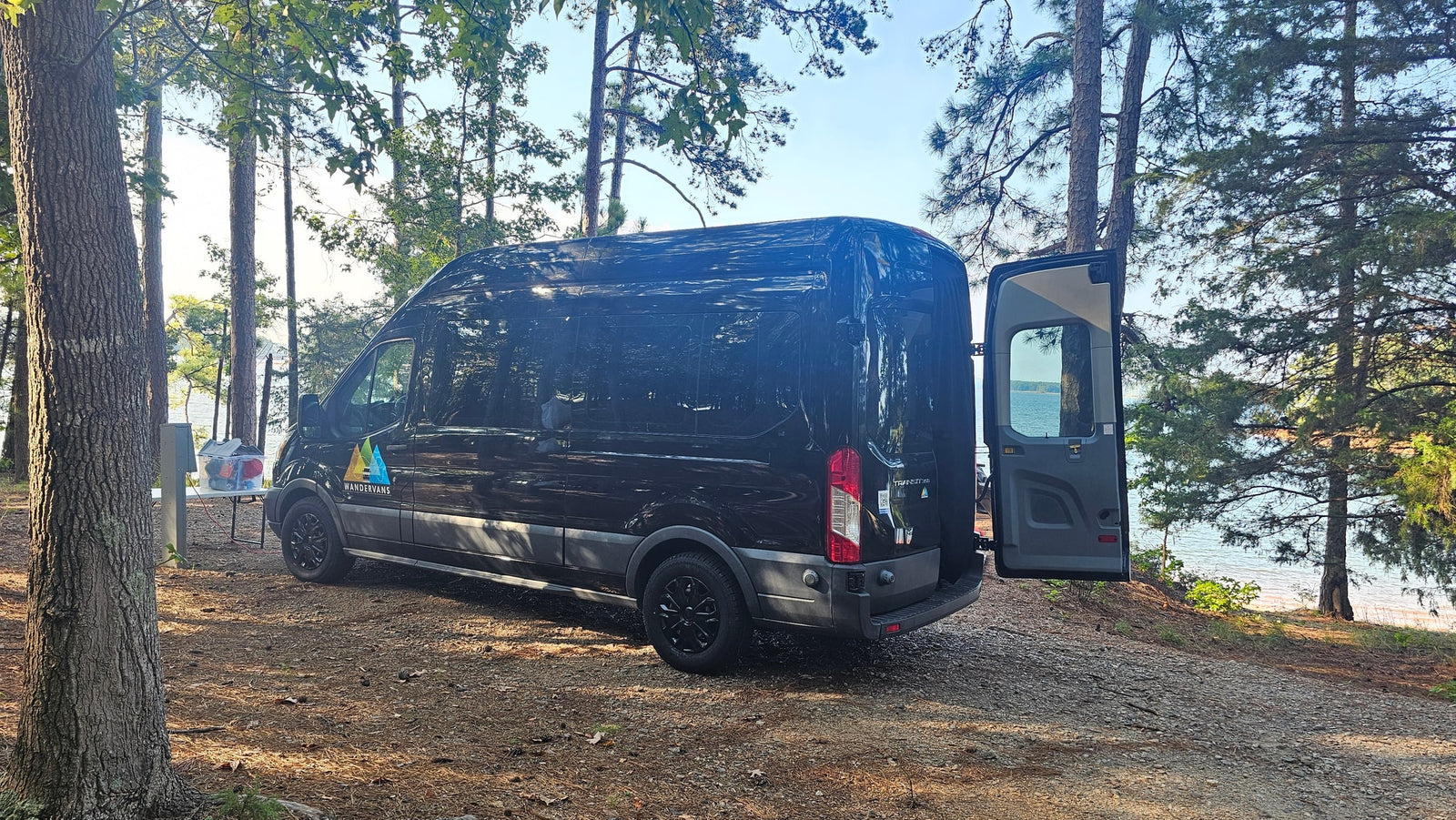 Black RV rental van at a shaded lakeside campsite. Rear doors open