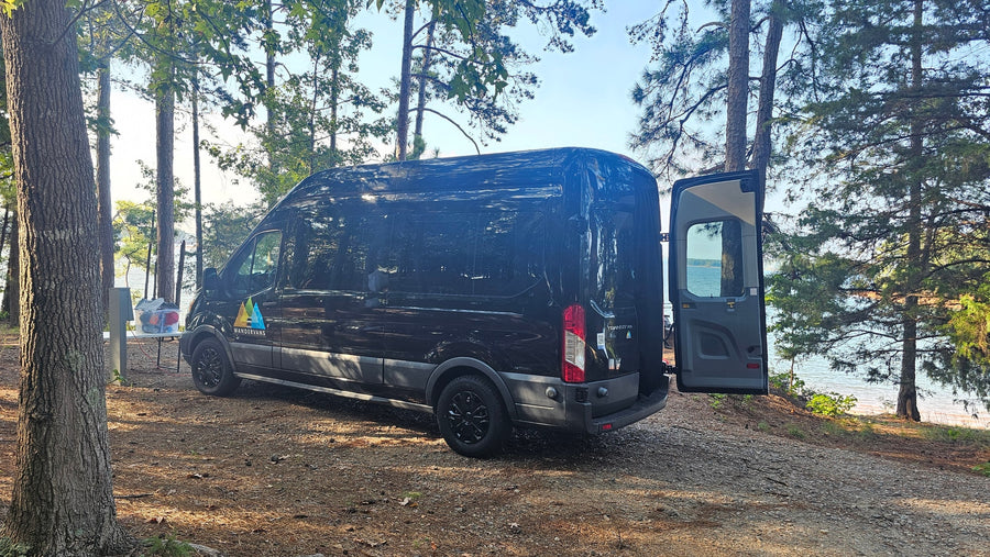 Black RV rental van at a shaded lakeside campsite. Rear doors open